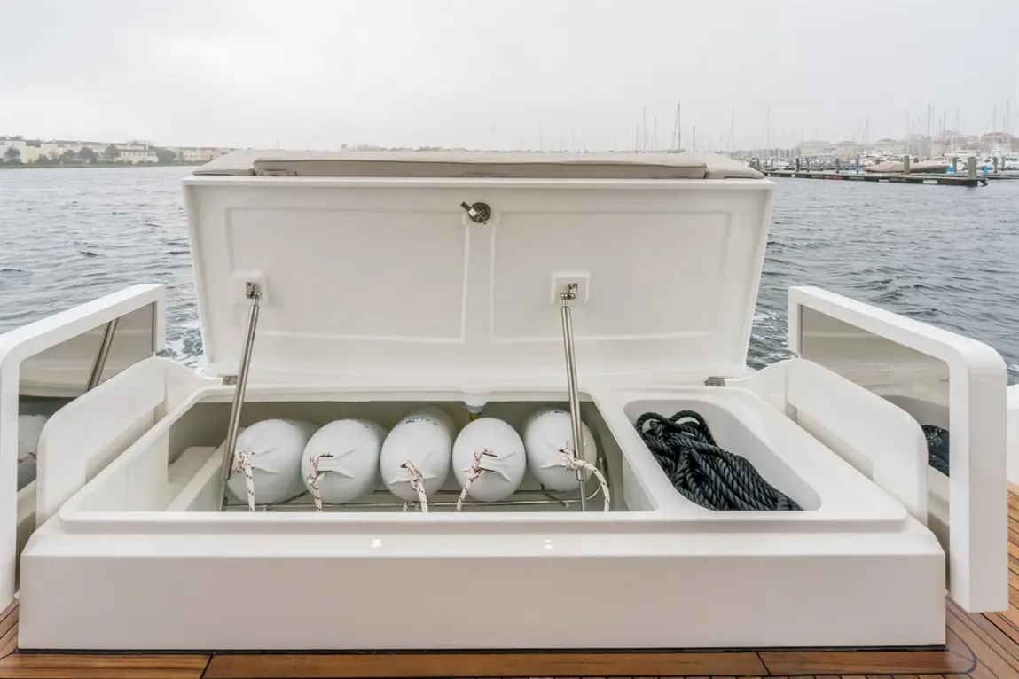 White boat storage compartment showing fenders and rope, with a view of a marina in the background on a cloudy day.